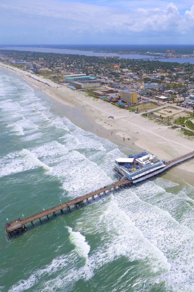 Aerial view of a coastal city with a long pier extending into the ocean, waves crashing along the sandy beach, and numerous buildings lining the shoreline under a partly cloudy sky.