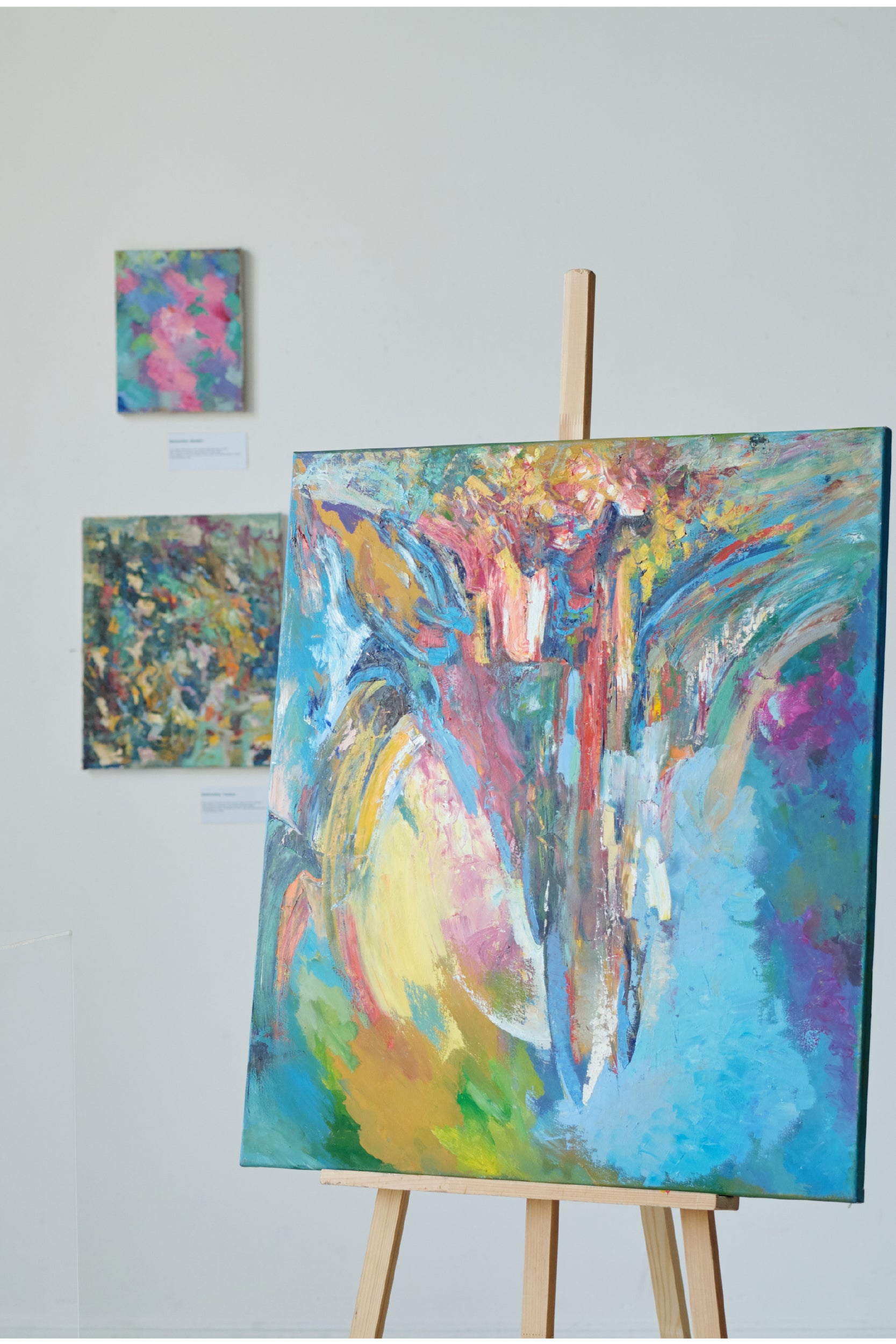 An abstract painting displayed on a wooden easel in an art gallery, featuring bold and vibrant colors including blue, pink, yellow, green, and red in sweeping, textured brushstrokes. Two additional abstract paintings are mounted on the white wall in the background, also composed of colorful, expressive patterns.