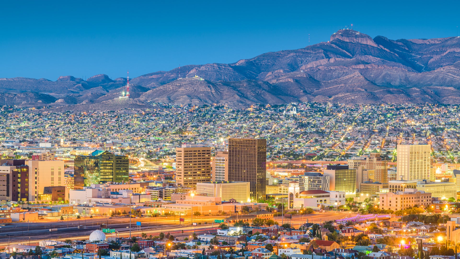 A vibrant evening view of El Paso, Texas, featuring the illuminated downtown skyline against a backdrop of rugged mountains. The cityscape extends into the distance with a mix of modern high-rise buildings and lower structures, while twinkling lights from residential areas add depth to the scene. The clear sky transitions into a deep blue, enhancing the contrast between the city and the natural landscape.