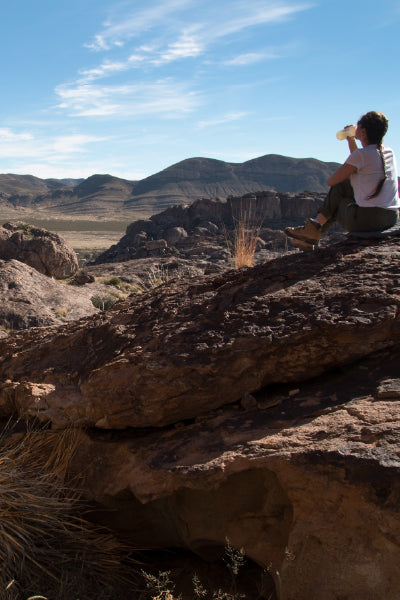 A person sits on a large rocky outcrop in a rugged desert landscape, sipping from a bottle while gazing into the distance. Rolling hills and mountains stretch across the horizon under a clear blue sky. The dry, rocky terrain is dotted with sparse vegetation, creating a serene and adventurous atmosphere.