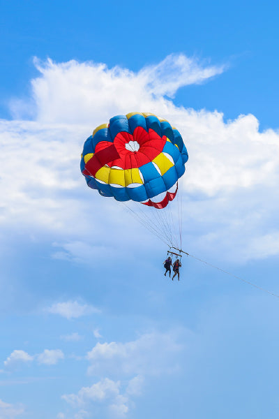 Two people are parasailing high in the sky, suspended beneath a vibrant multicolored parachute with red, yellow, and blue sections. They are harnessed to a metal bar, with a taut rope extending downward, likely connecting them to a boat. The backdrop features a bright blue sky with scattered white clouds.