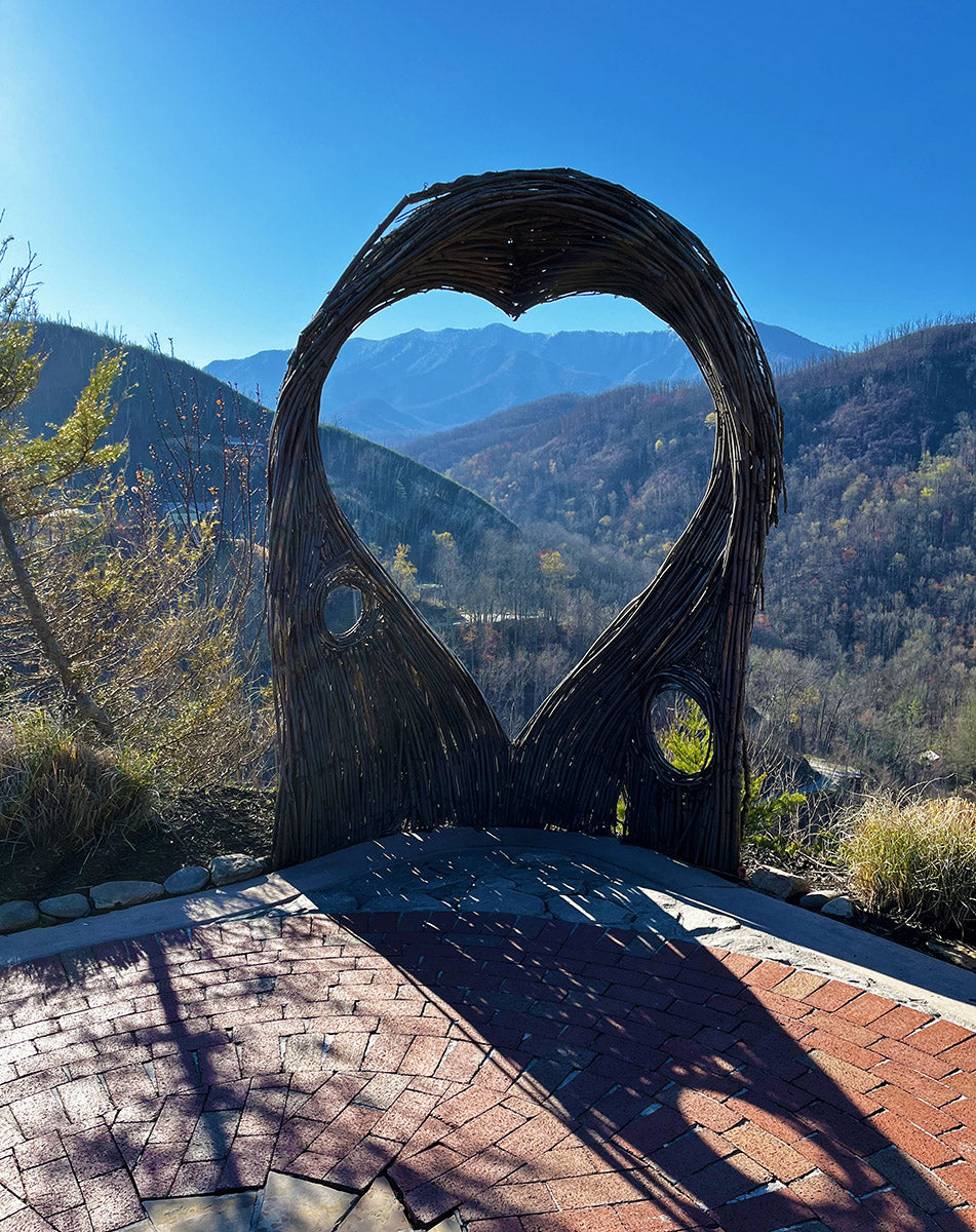 A scenic mountain view framed by a heart-shaped wooden sculpture made of intertwined branches. The natural archway stands on a brick-paved platform, casting long shadows in the sunlight. The background showcases rolling mountains under a clear blue sky, creating a picturesque and romantic setting.