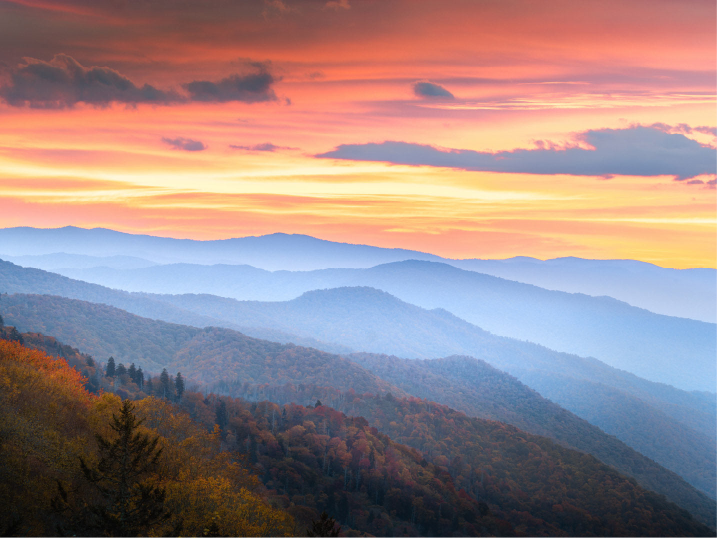 A breathtaking view of rolling mountains covered in autumn foliage, with layers of blue ridges fading into the distance. The sky is illuminated by a vibrant sunset, casting warm hues of orange, pink, and yellow across scattered clouds. The scene evokes a sense of tranquility and natural beauty.