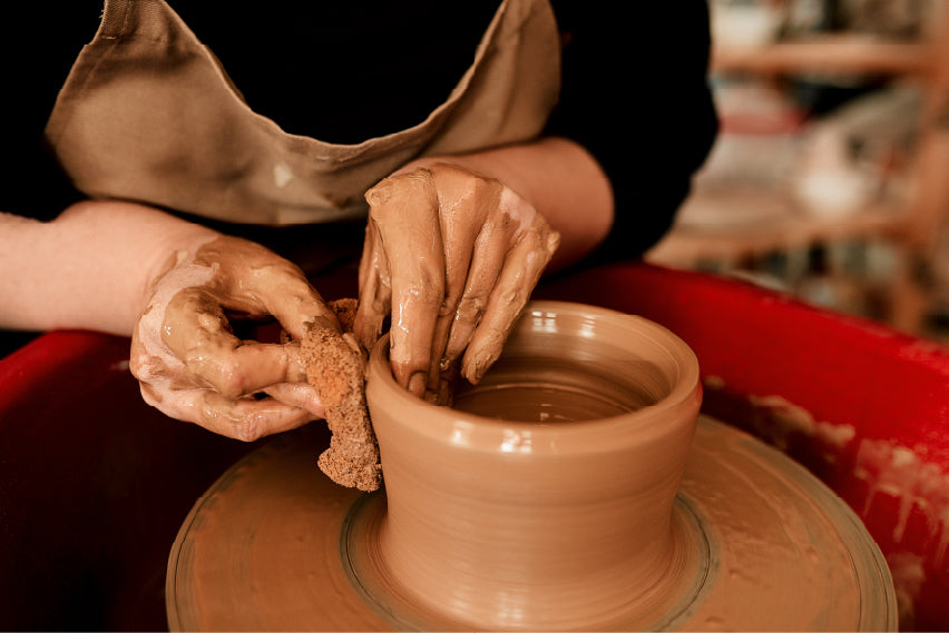 A person shaping a clay pot on a pottery wheel, with hands covered in wet clay. One hand is using a sponge to smooth the surface while the other hand supports and forms the pot's structure. The background suggests a pottery studio with shelves and tools.