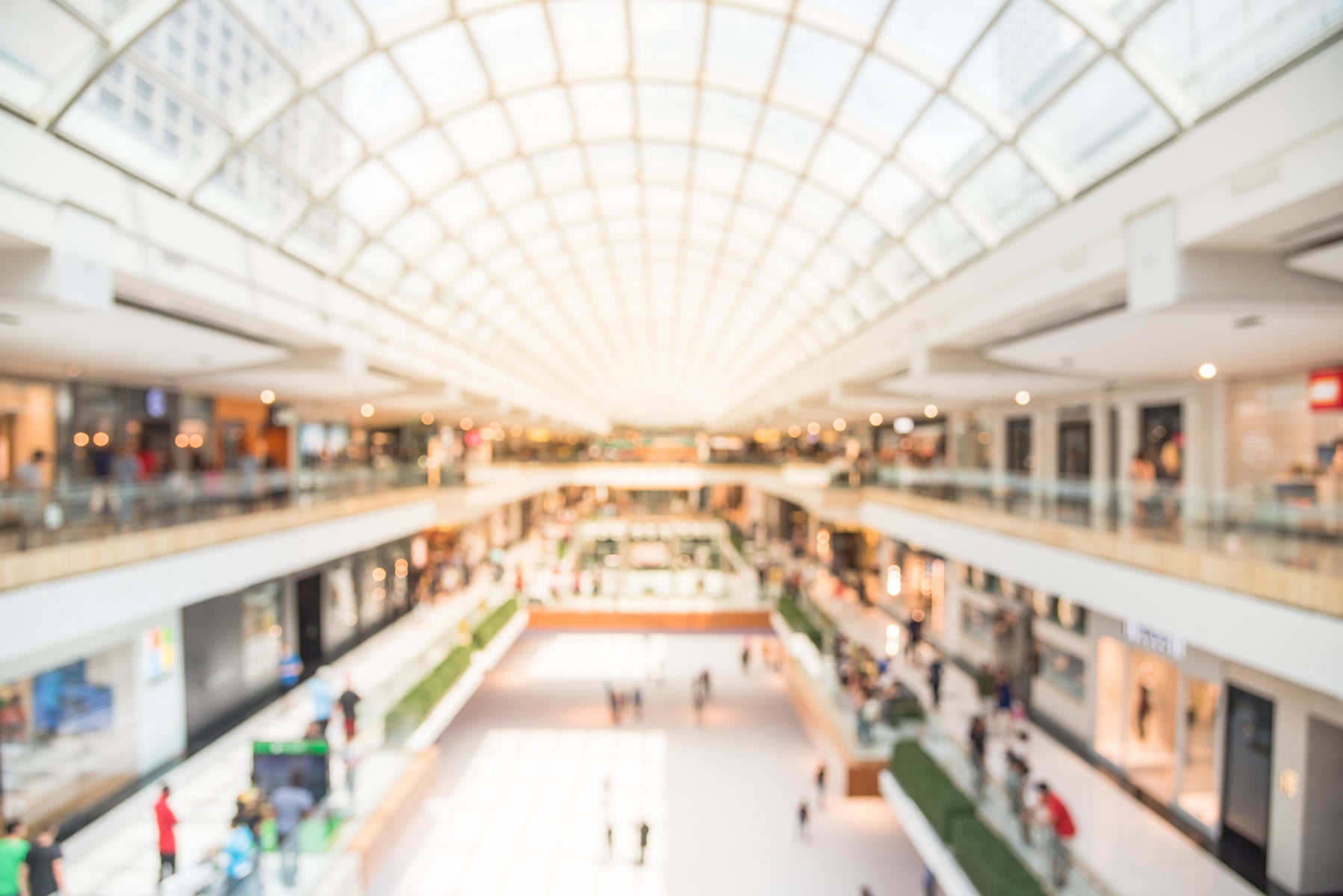 A blurred image of a modern shopping mall with multiple levels, a glass-domed ceiling, and bright lighting. Shoppers walk along the corridors and browse stores, creating a lively atmosphere.