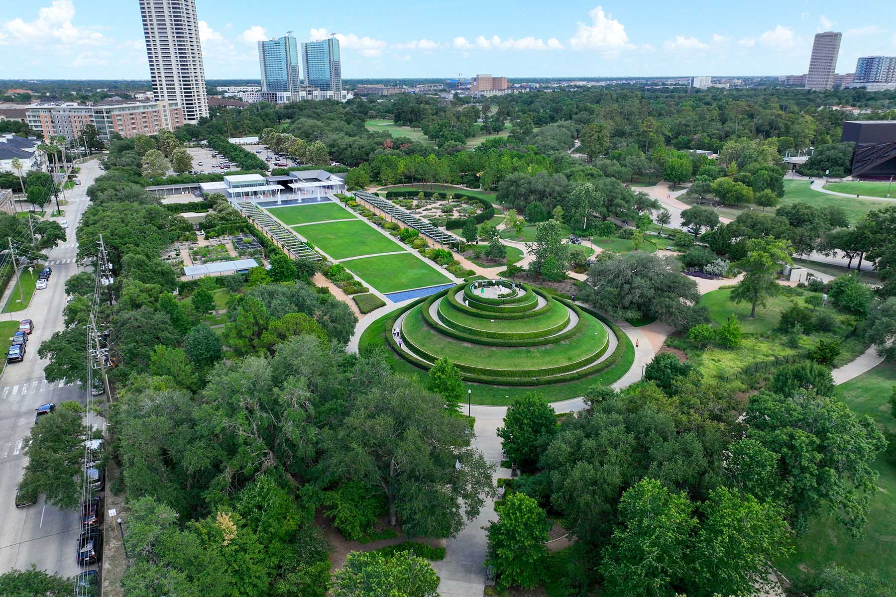 An aerial view of a lush urban park featuring a distinctive circular tiered garden at its center. The park is surrounded by trees, walking paths, and a modern building with green lawns. In the background, high-rise buildings and a cityscape are visible.