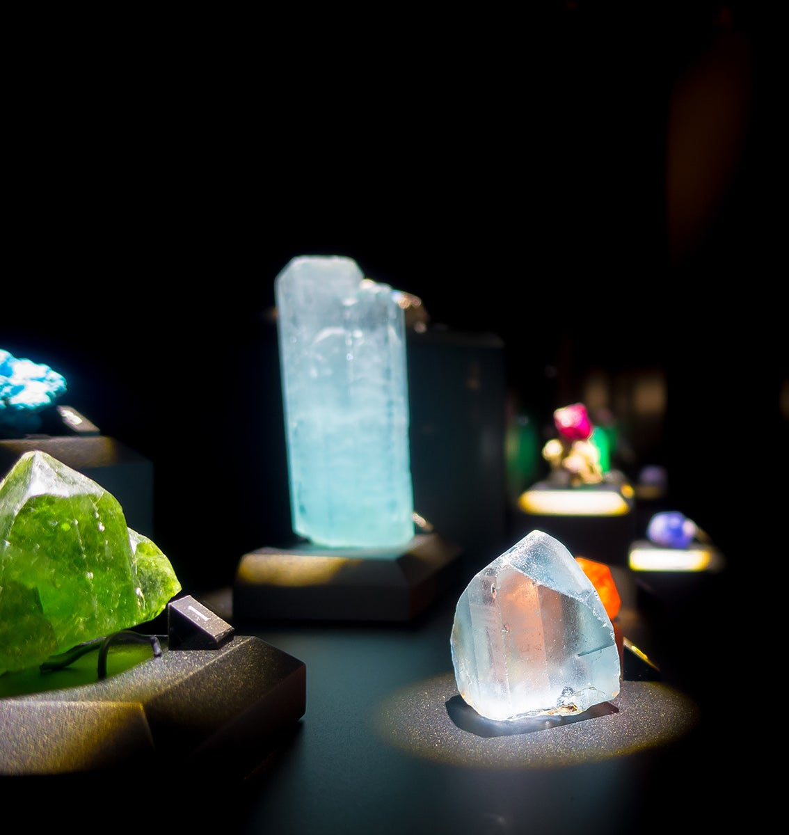 A display of colorful gemstones and crystals illuminated against a dark background. The focus is on a translucent blue crystal in the foreground, with other vibrant minerals, including green and orange stones, arranged on pedestals in the distance.