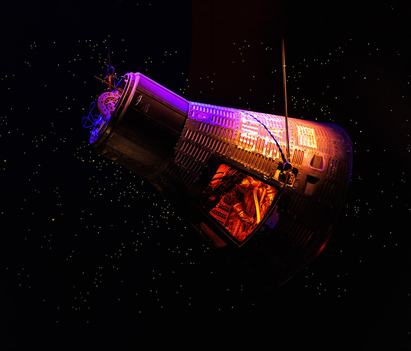 A historic space capsule suspended in a dark exhibit, illuminated by purple and orange lighting. The capsule’s metallic exterior features intricate paneling, and its window reveals a detailed interior. The backdrop simulates outer space with small glowing stars.