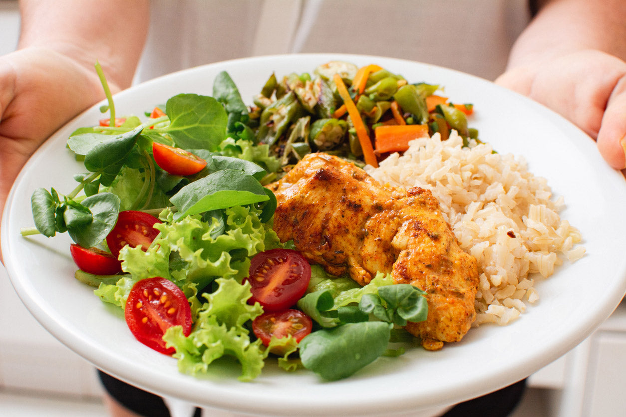 A close-up of a plate filled with a balanced meal, including a fresh green salad with cherry tomatoes, seasoned grilled chicken, a side of brown rice, and stir-fried vegetables such as carrots and bell peppers. The plate is being held by a person, with their hands visible in the background, offering a healthy and colorful dish.