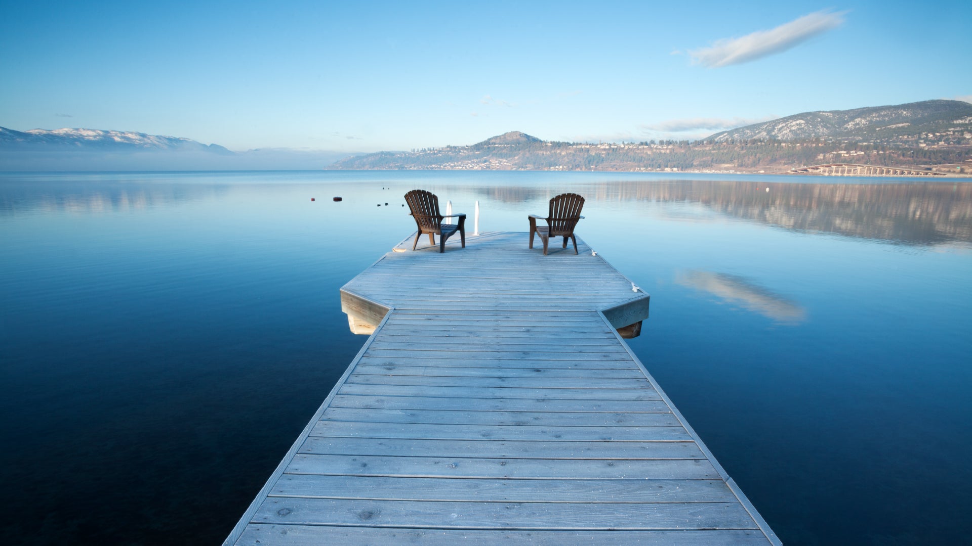 A tranquil lakeside scene featuring a wooden dock extending into the still, reflective waters. Two Adirondack chairs sit at the end of the dock, facing the serene lake, inviting relaxation. In the background, distant mountains with snow-capped peaks stretch across the horizon under a soft blue sky with wispy clouds. The calm water mirrors the sky and surrounding landscape, enhancing the peaceful ambiance of the setting.