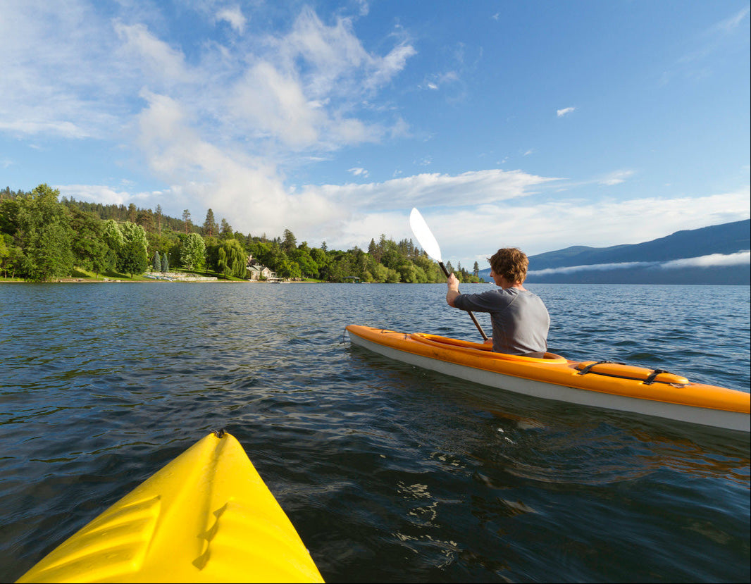 A scenic view from a yellow kayak on a calm lake, looking toward another person paddling a bright orange kayak. The kayaker is wearing a gray shirt and is actively rowing with a white paddle. The background features a lush, tree-lined shore with houses and a distant mountain range partially covered in clouds. The sky is blue with scattered white clouds, creating a peaceful and inviting atmosphere.