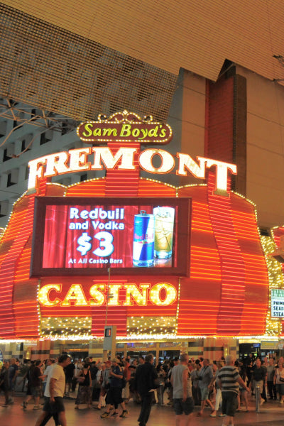Nighttime view of the entrance to Sam Boyd's Fremont Casino in Las Vegas, featuring bright red and yellow neon lights. A large digital sign above the entrance advertises a \$3 Redbull and vodka drink special. The street in front of the casino is crowded with people walking and gathering under the lit-up canopy of Fremont Street Experience.
