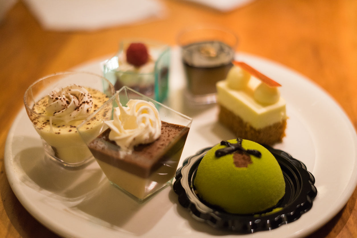 A plate of assorted miniature desserts arranged on a white dish, featuring a variety of textures and flavors. The selection includes creamy custards topped with whipped cream and cocoa powder, a layered chocolate and vanilla mousse in a glass, a small cheesecake with decorative toppings, and a vibrant green dome-shaped pastry with chocolate accents on a black tray. The desserts are beautifully presented, showcasing intricate details and inviting colors against a softly blurred background.