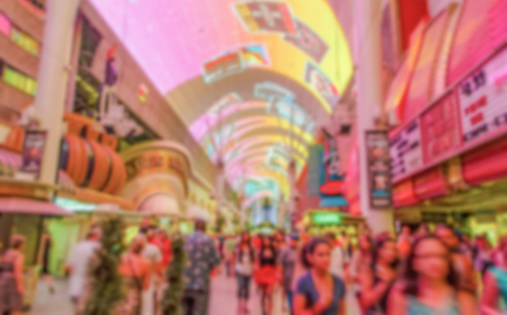 A bustling, colorful scene of a crowded pedestrian street with a bright, illuminated canopy overhead, displaying vibrant digital graphics and signs. The surrounding buildings are adorned with bold, neon lights and marquee advertisements, creating a lively, festive atmosphere. The image has a blurred effect, enhancing the feeling of motion and excitement.