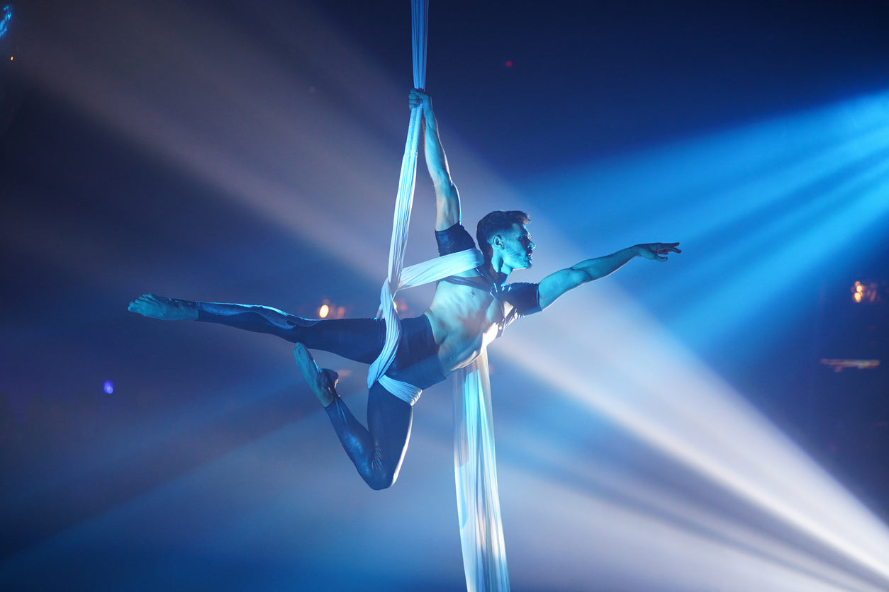 A male aerial silk performer suspended mid-air, gracefully balancing with one arm extended and his body wrapped in white silks. He wears a sleek black and silver costume, and dramatic blue stage lighting casts beams of light across the darkened background, creating a mesmerizing and theatrical atmosphere.