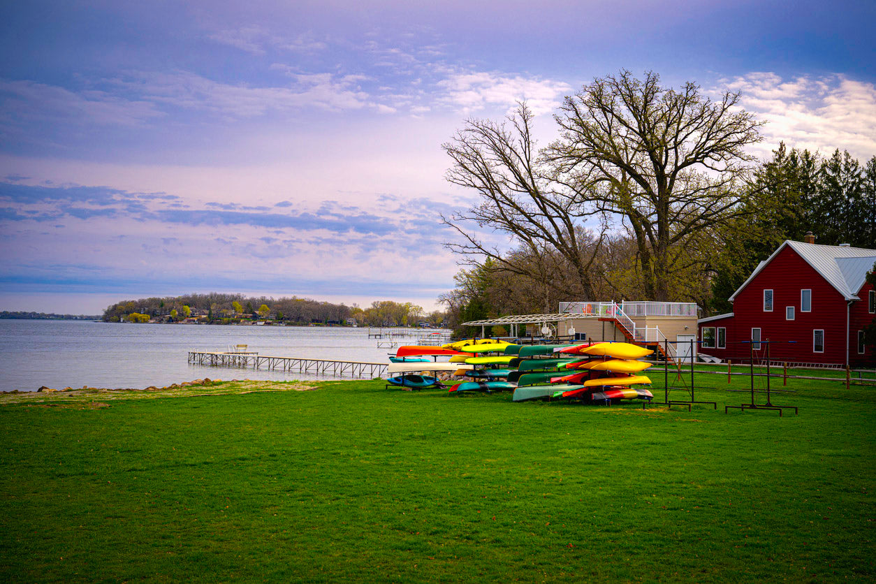 A peaceful lakeside scene featuring a grassy shoreline with a rack of colorful kayaks and canoes neatly stacked. A wooden dock extends into the calm water, with a few houses visible in the distance along the opposite shore. To the right, a red barn-like building with a white roof stands among tall, leafless trees, suggesting early spring or late autumn. The sky is a mix of soft blue and gray hues, with scattered clouds adding depth to the tranquil setting.