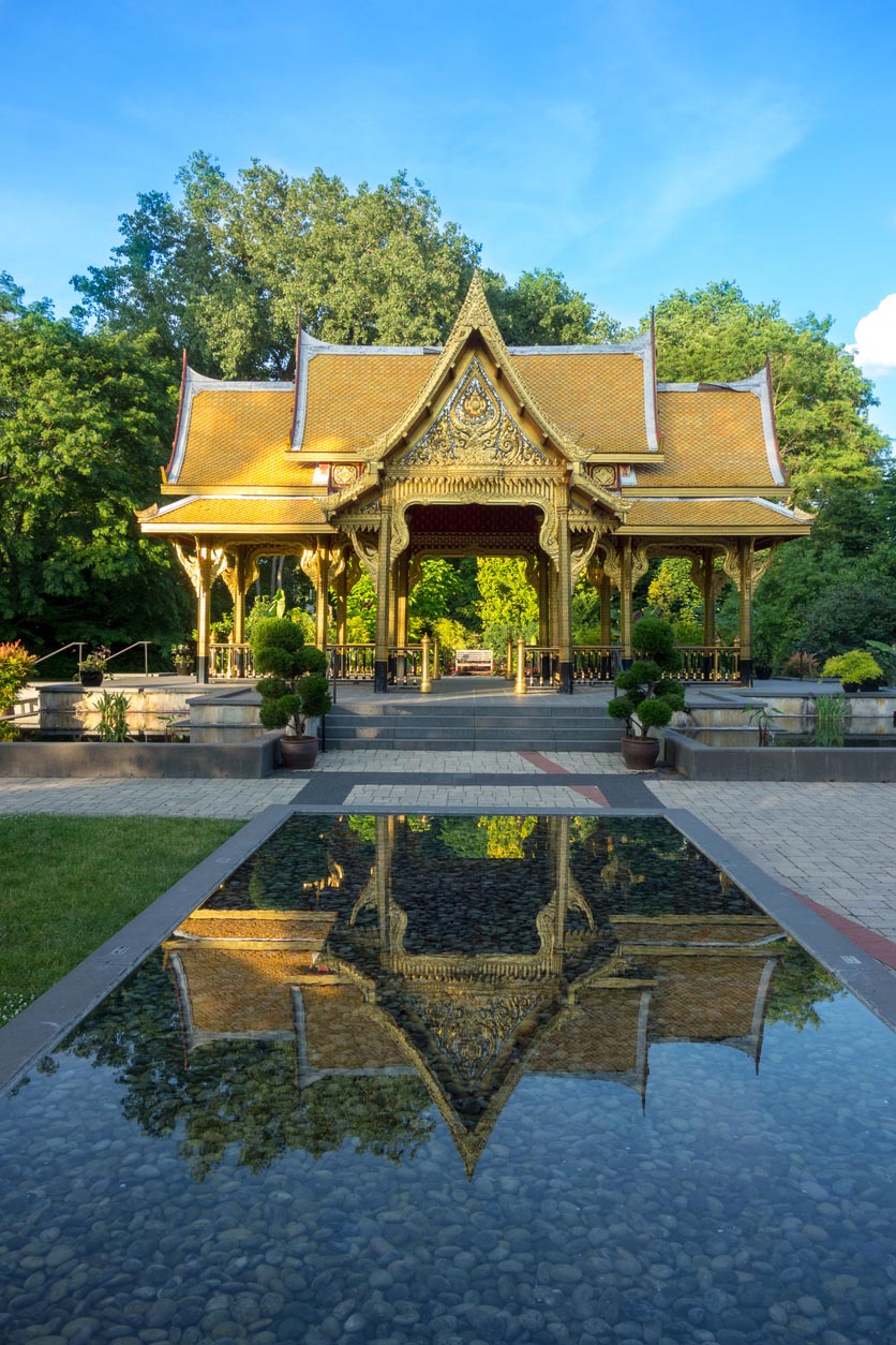 A traditional Thai pavilion with intricate gold detailing and a tiered roof stands in a lush park, surrounded by greenery. A reflecting pool in the foreground creates a symmetrical mirror image of the ornate structure, with smooth stones visible beneath the water. The scene is bathed in warm sunlight, highlighting the vibrant colors and craftsmanship.