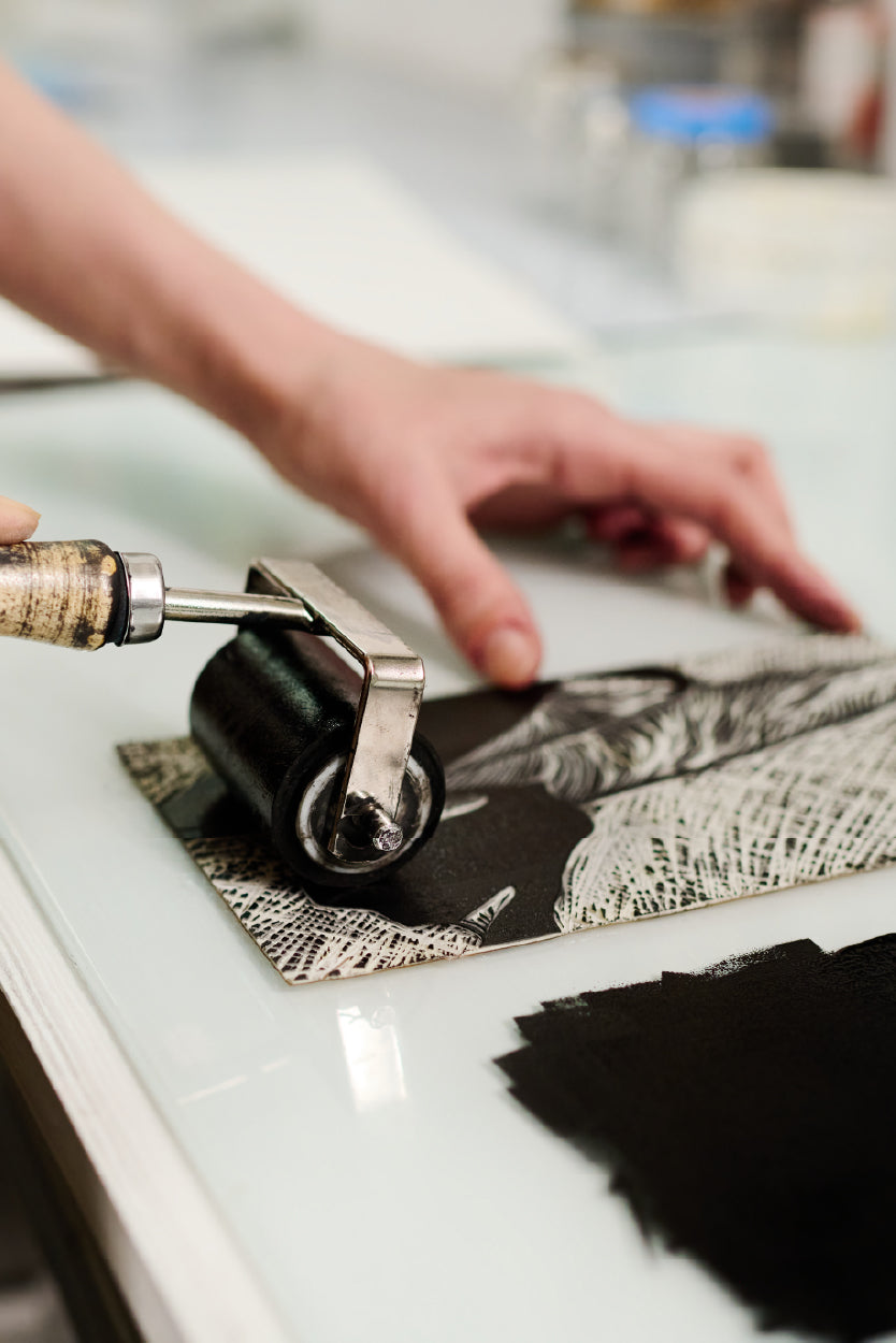 A close-up of a person using a brayer roller to apply black ink onto a carved linoleum block, preparing it for printmaking. The detailed carving on the block features intricate patterns, and the person's hand carefully holds it steady on a clean, white surface. The scene captures the artistic process of creating a linocut print.