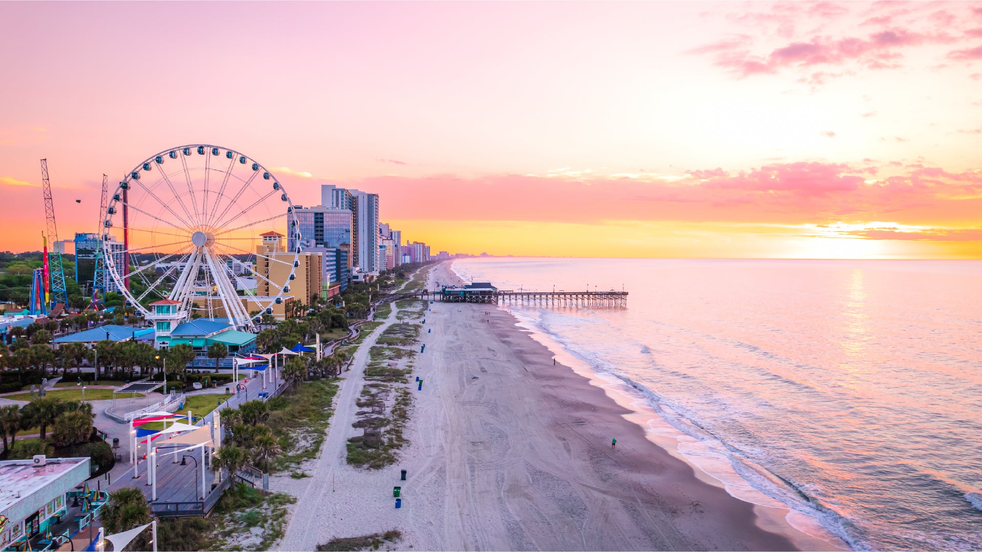 A stunning sunset over Myrtle Beach, South Carolina, with the sky painted in shades of pink, orange, and purple. The Ferris wheel stands tall near the bustling boardwalk, while a wooden pier stretches into the calm ocean. High-rise hotels and colorful attractions line the coast, and the sandy beach, dotted with a few visitors, meets the gently rolling waves.