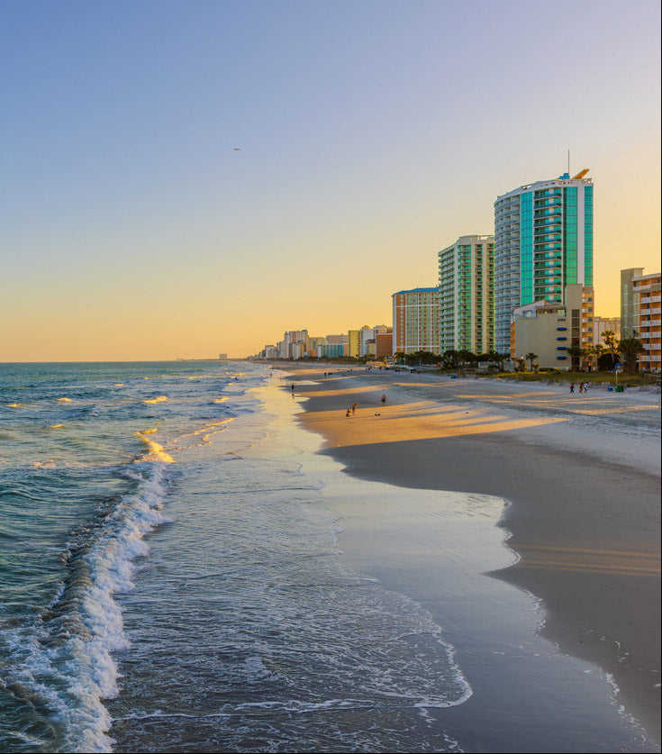 A scenic view of a beach at sunset, with gentle ocean waves rolling onto the sandy shore. The golden sunlight reflects off the water, creating a warm and inviting atmosphere. In the background, a row of modern high-rise buildings lines the coastline, contrasting with the natural beauty of the ocean. Few people can be seen walking along the shoreline, enjoying the peaceful ambiance of the evening.