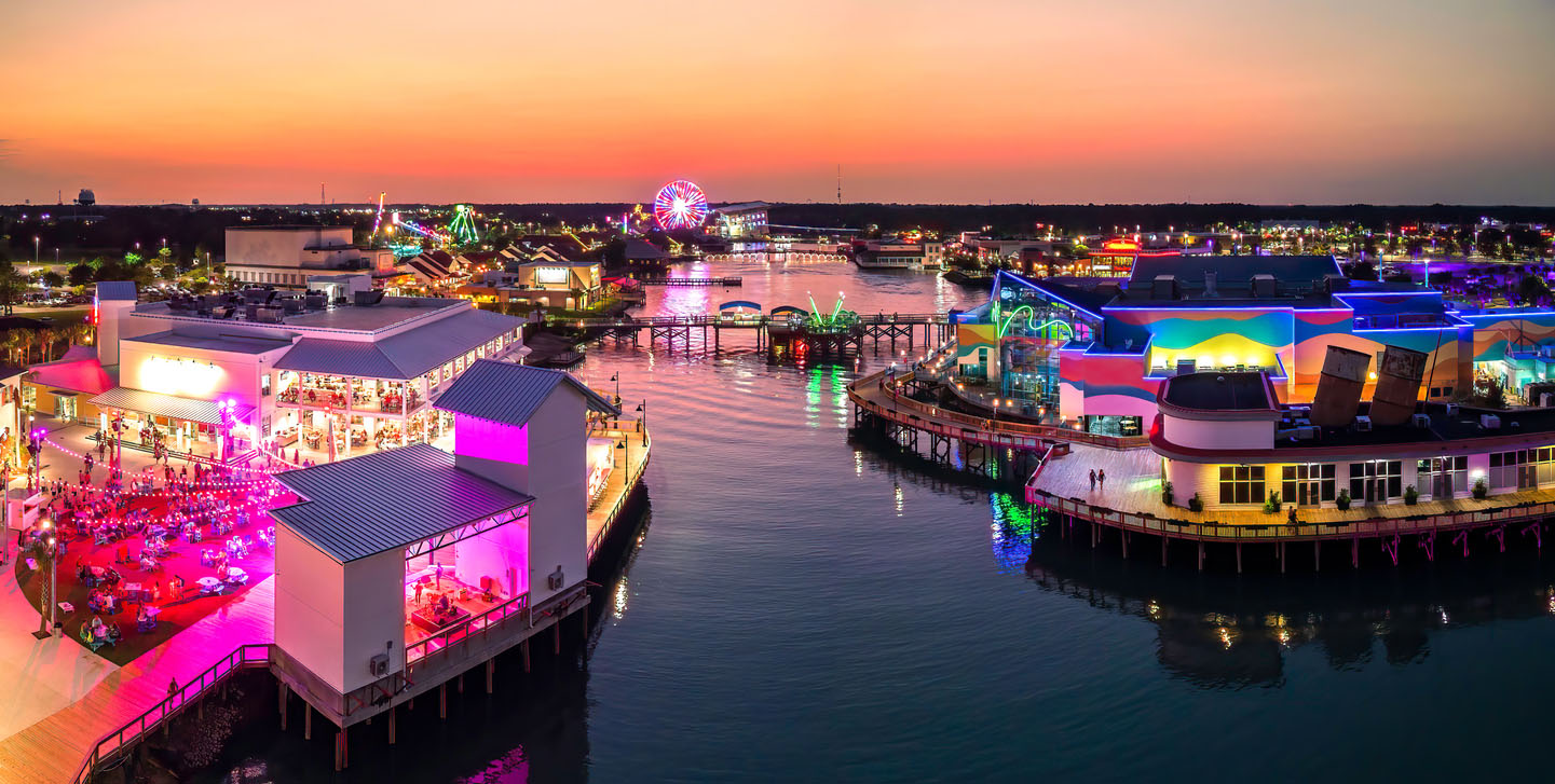 A vibrant waterfront entertainment district illuminated by colorful lights at sunset. The scene features modern buildings, open-air dining areas, and boardwalks extending over calm waters. A Ferris wheel glows in the distance, along with other amusement attractions, reflecting on the water. The lively atmosphere is enhanced by bright neon and LED lights decorating the structures, creating a visually stunning nighttime setting.