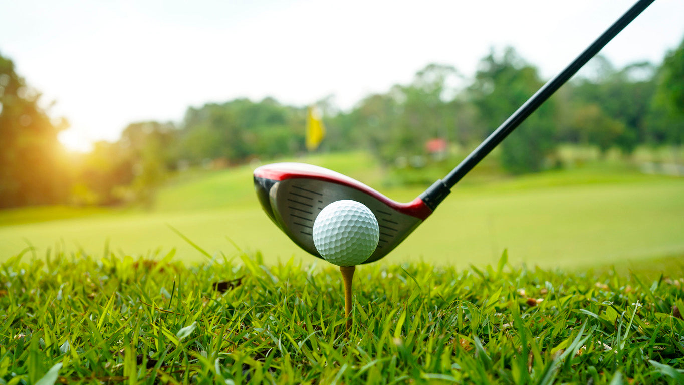 A close-up photograph of a golf driver positioned behind a golf ball on a tee, ready for a shot. The scene is set on a lush green golf course with the fairway and a yellow flag visible in the blurred background. The sunlight casts a warm glow, enhancing the vibrant colors of the grass and trees.