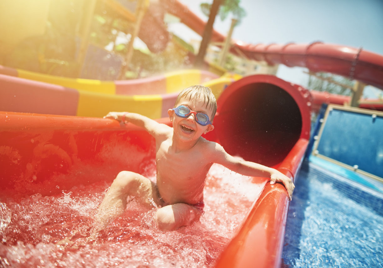 A young boy wearing blue goggles is joyfully sliding down a bright red water slide at a water park. His wet hair and wide smile reflect excitement as he splashes into the pool at the bottom. Sunlight shines brightly, creating a warm and vibrant atmosphere. In the background, colorful slides and water attractions are visible, adding to the fun-filled scene.