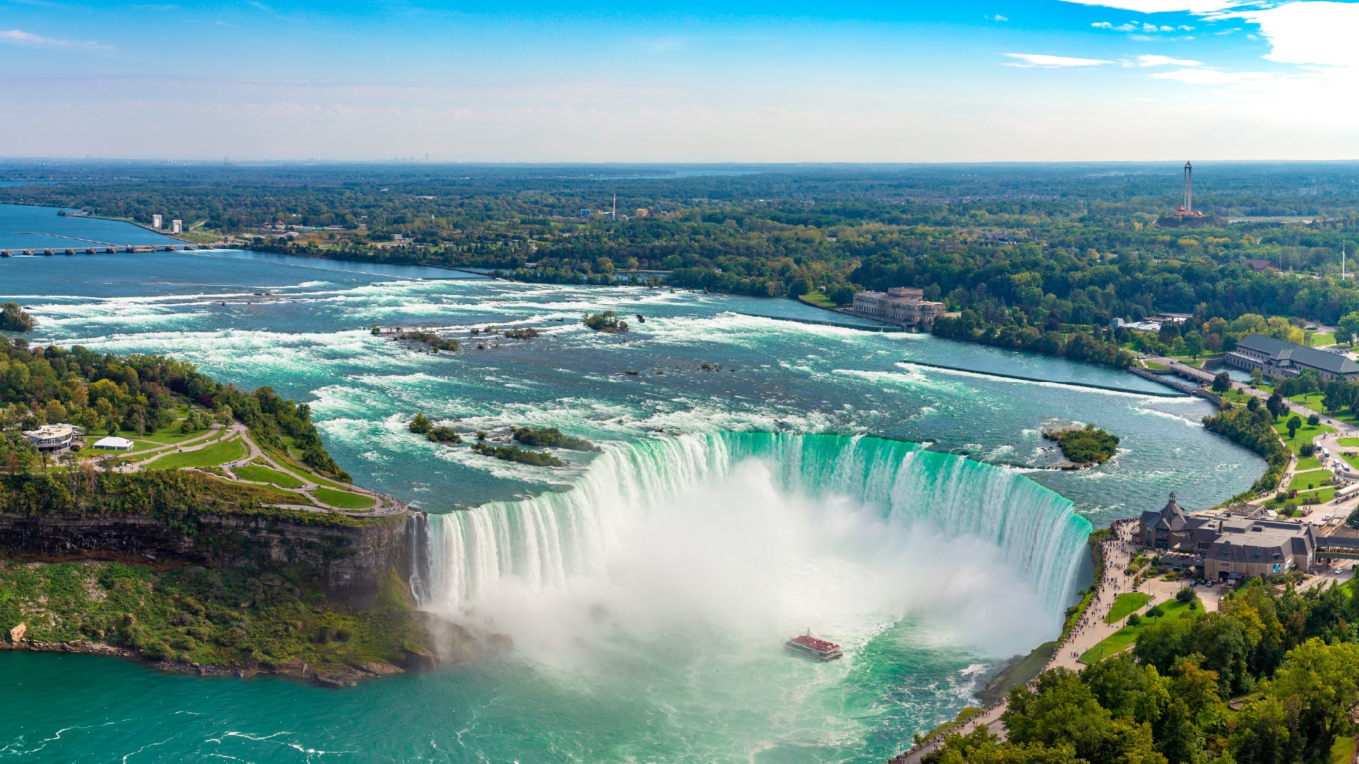 A breathtaking aerial view of Niagara Falls, showcasing the powerful Horseshoe Falls with turquoise water cascading over the edge, creating a misty cloud. A tour boat navigates near the falls, giving a sense of scale to the immense waterfall. Surrounding the falls is lush greenery, walkways, and observation areas with visitors admiring the natural wonder. In the background, a river with rapids flows toward the falls, and a bridge spans across the water. The sky is bright blue with scattered clouds.