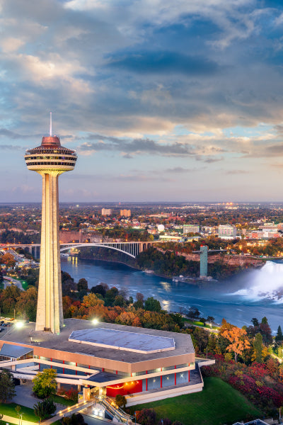 A scenic view of the Skylon Tower in Niagara Falls, Canada, captured at sunset. The tall observation tower stands prominently in the foreground, overlooking the Niagara River and the Rainbow Bridge, which connects Canada and the United States. The Horseshoe Falls can be seen in the background, partially shrouded in mist. The cityscape extends into the horizon, framed by a colorful autumn landscape and a dramatic, partly cloudy sky.