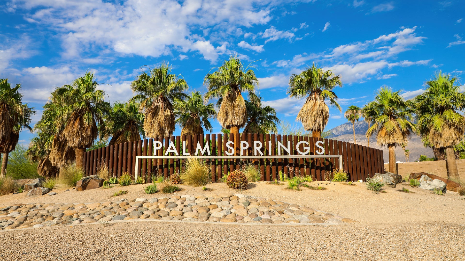 A vibrant and welcoming entrance sign to Palm Springs, California, set against a backdrop of tall palm trees and a bright blue sky with scattered clouds. The sign features bold white lettering on a rustic wooden fence, surrounded by desert landscaping with rocks, sand, and drought-resistant plants. In the distance, mountains rise, adding to the picturesque beauty of this famous desert resort city.