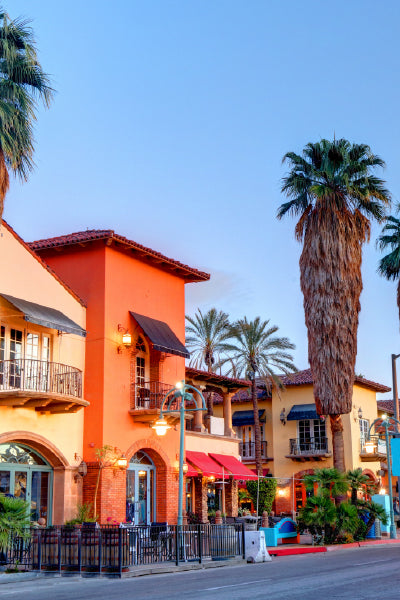 A vibrant street scene at sunset featuring colorful Spanish-style buildings with arched windows, wrought-iron balconies, and red-tile roofs. Tall palm trees line the street, and outdoor seating areas hint at cafés or restaurants welcoming visitors. The clear sky and warm lighting create a relaxed, inviting atmosphere.