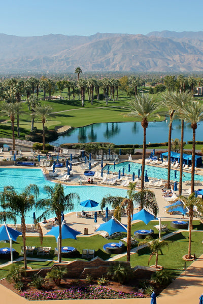 A luxurious resort scene featuring multiple swimming pools surrounded by blue umbrellas and lounge chairs, set amid lush green palm trees. In the background, there is a tranquil lake, a manicured golf course, and a range of arid mountains under a clear blue sky.