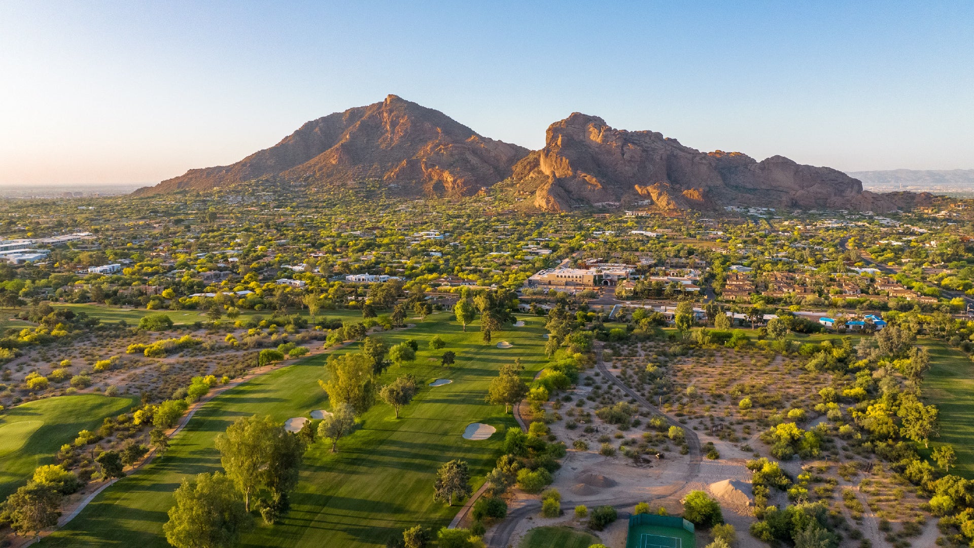 A breathtaking aerial view of Paradise Valley, Arizona, with Camelback Mountain dominating the background. The rugged peaks of the mountain are bathed in warm sunlight, contrasting against the lush greenery of a golf course in the foreground. The surrounding desert landscape is dotted with luxurious homes, resorts, and vibrant vegetation, creating a picturesque blend of nature and urban development. The clear blue sky stretches above, enhancing the beauty of this scenic southwestern destination.