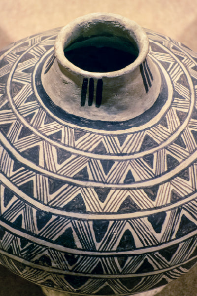 Close-up of a traditional Native American ceramic pot featuring intricate black geometric patterns, including zigzags and triangles, painted on a beige surface with a narrow neck and three vertical black lines near the rim.