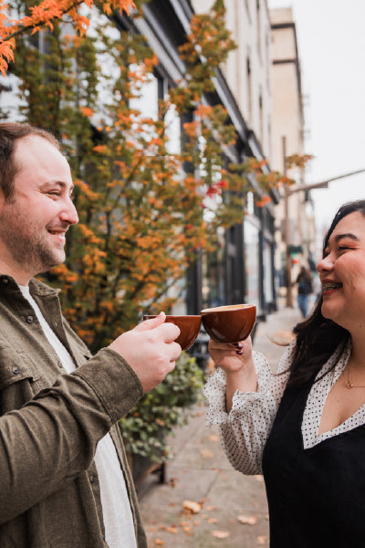 A smiling man and woman stand outside on a city sidewalk, holding brown coffee mugs and toasting each other. Autumn leaves on a nearby tree add warm color to the scene. The background shows storefronts and buildings along the street, suggesting a cheerful urban coffee outing.