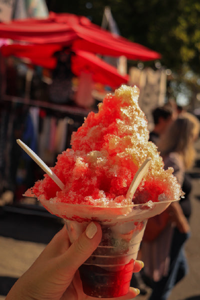 Close-up of a hand holding a colorful shaved ice dessert in a clear plastic cup with red and brown syrup, two white spoons sticking out, and a vibrant outdoor market scene with red umbrellas and blurred people in the background.