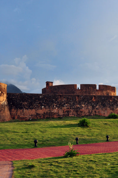 Historic stone fortress with thick, weathered walls and battlements, situated on a grassy hill with a red brick walkway in the foreground under a partly cloudy sky.