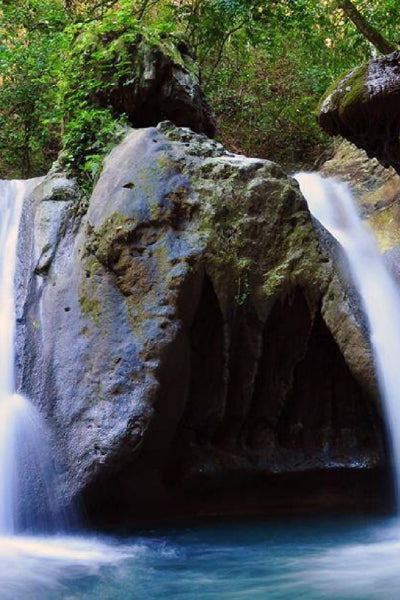 Scenic waterfall flowing gently over large moss-covered rocks into a clear blue pool, surrounded by lush green foliage in a forest setting.