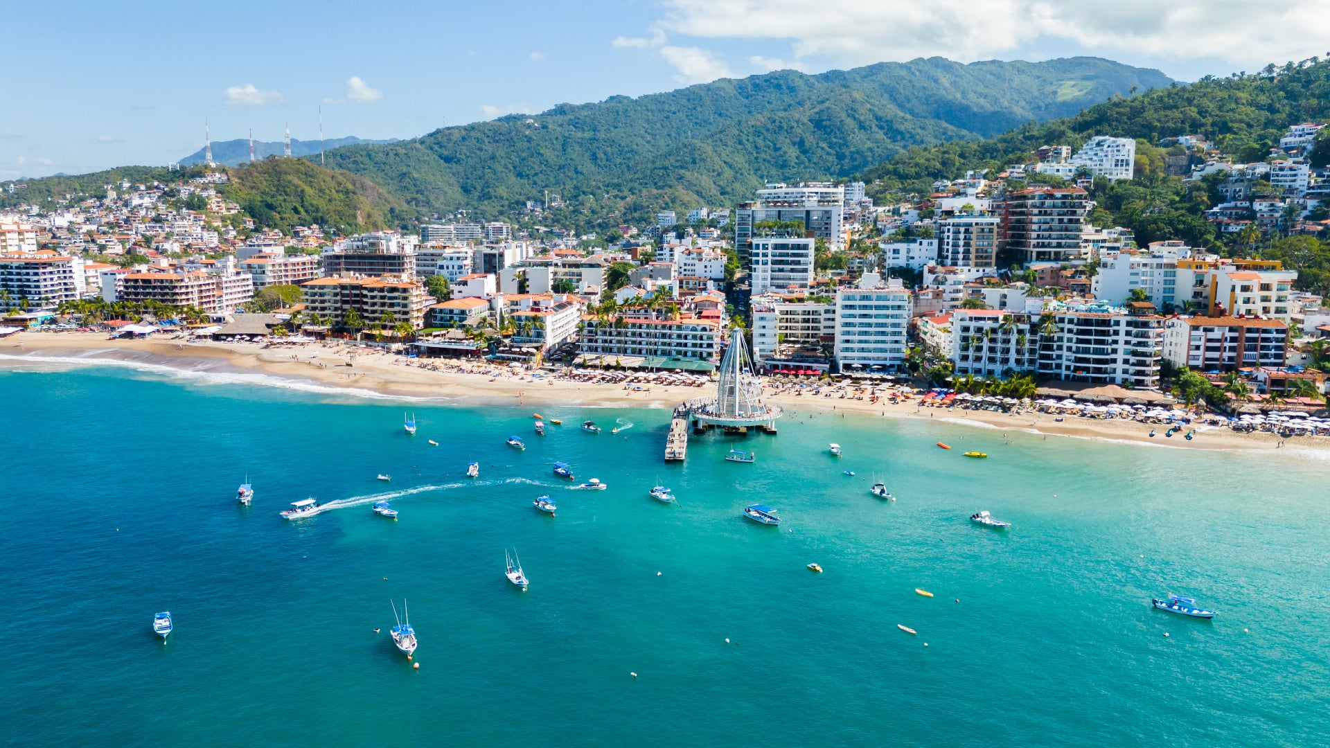 A scenic aerial view of a tropical beachside city with turquoise waters, a pier extending into the ocean, and boats scattered across the sea. The shoreline is lined with high-rise hotels, resorts, and residential buildings, with lush green mountains in the background. The beach is dotted with umbrellas and sunbathers, creating a vibrant and inviting coastal scene.