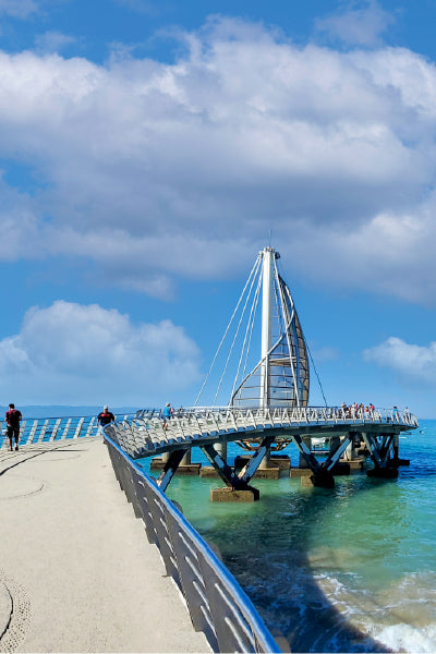 A modern pier extends over turquoise ocean water, leading to a circular observation platform with a striking sail-shaped metal structure at the center. People are walking along the curved path and enjoying the scenic view under a bright blue sky with fluffy clouds. The pier features sleek metal railings and stands on sturdy concrete supports above the waves.