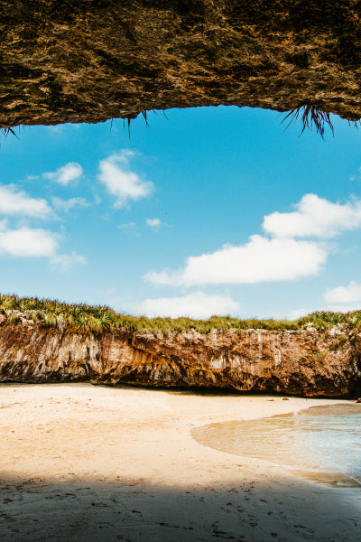 A hidden beach surrounded by rocky cliffs with an open skylight above, revealing a clear blue sky dotted with fluffy white clouds. The sandy shore and gentle waves create a peaceful atmosphere, while the grass-covered edges of the rocky enclosure give the area a secluded, natural charm.