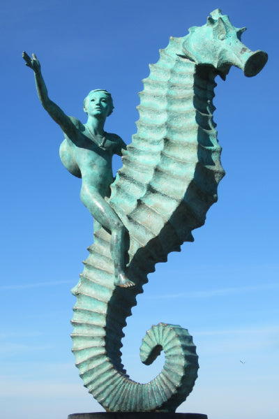 A bronze statue of a young boy riding a giant seahorse, with one arm raised in the air as if waving or reaching out. The sculpture is set against a clear blue sky, giving it a dramatic and uplifting appearance. The detailed texture of the seahorse and the boy’s expression add a sense of movement and wonder to the scene.