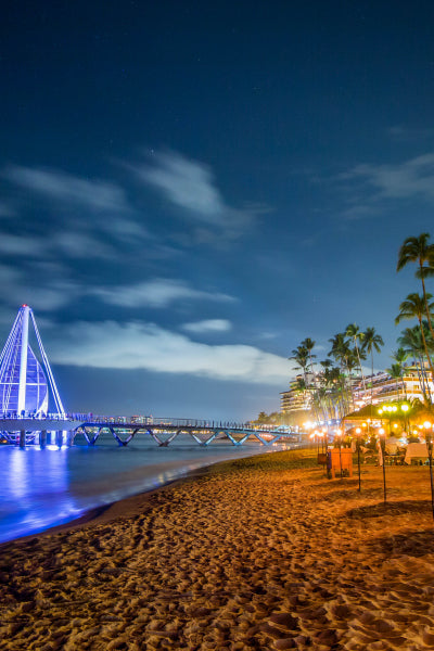 A night view of a beach with a well-lit modern pier extending into the ocean, featuring a distinctive sail-like structure illuminated in blue. The sandy shore is lined with palm trees and warmly lit outdoor dining tables, creating a vibrant, inviting atmosphere. In the background, resort buildings glow against the night sky filled with scattered clouds.