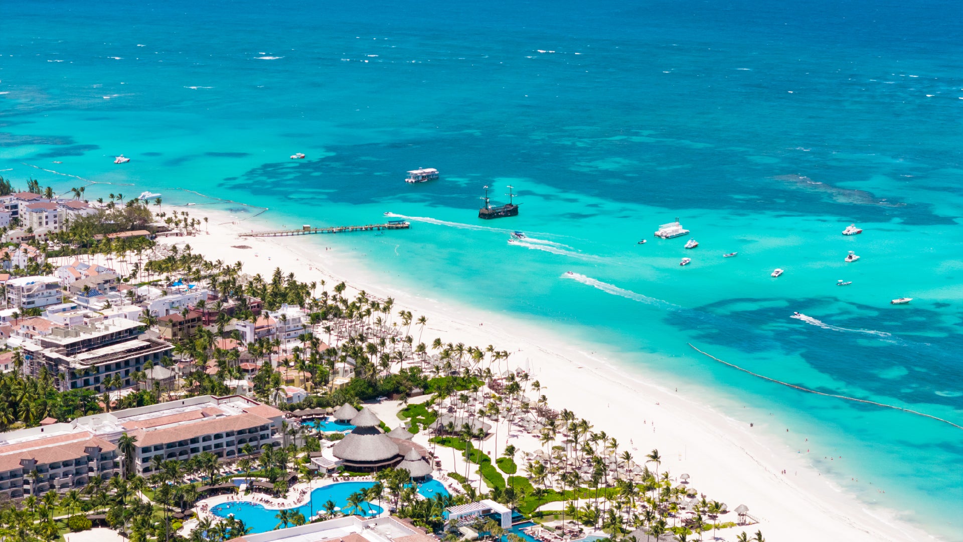 A breathtaking aerial view of a tropical beach resort with white sandy shores, turquoise waters, and lush palm trees. The scene features luxurious resorts with pools, a wooden pier extending into the ocean, and boats, including a pirate-style ship, sailing across the vibrant sea. The crystal-clear water showcases varying shades of blue, indicating coral reefs and sandbars beneath the surface.