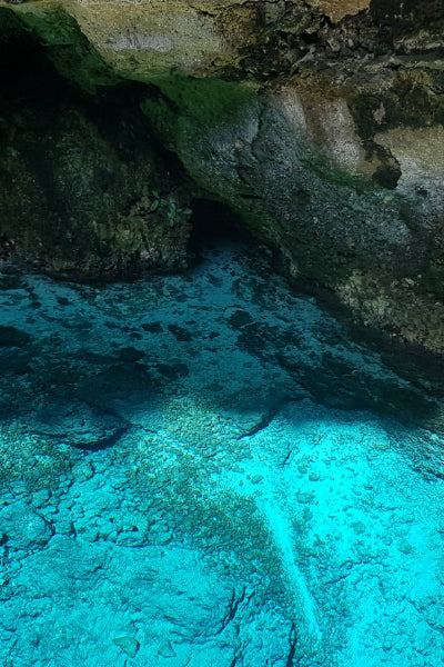 A clear blue water cave with rocky formations and a dark entrance in the background. The shallow water reveals the rocky seabed beneath, creating a vivid contrast between the bright turquoise water and the shadowy cave interior.