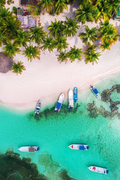 Aerial view of a tropical beach with white sand and lush green palm trees lining the shore. Several small boats are anchored in the clear turquoise water near the beach, creating a vibrant and serene coastal scene.