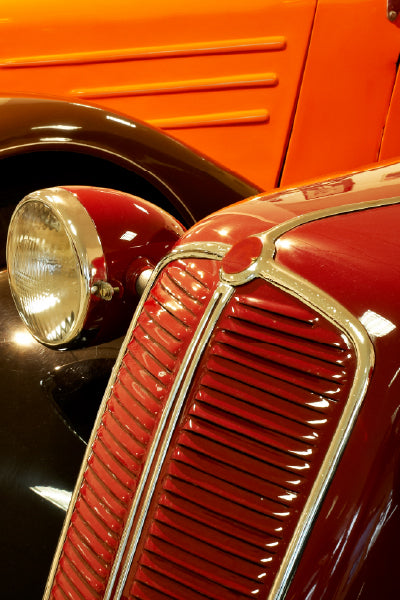 Close-up view of the front sections of two vintage cars, one painted glossy red and the other bright orange. The image highlights the red car’s grille and round headlight, as well as part of the orange car's fender and side panel. The colors are vibrant, and the chrome details reflect the light.