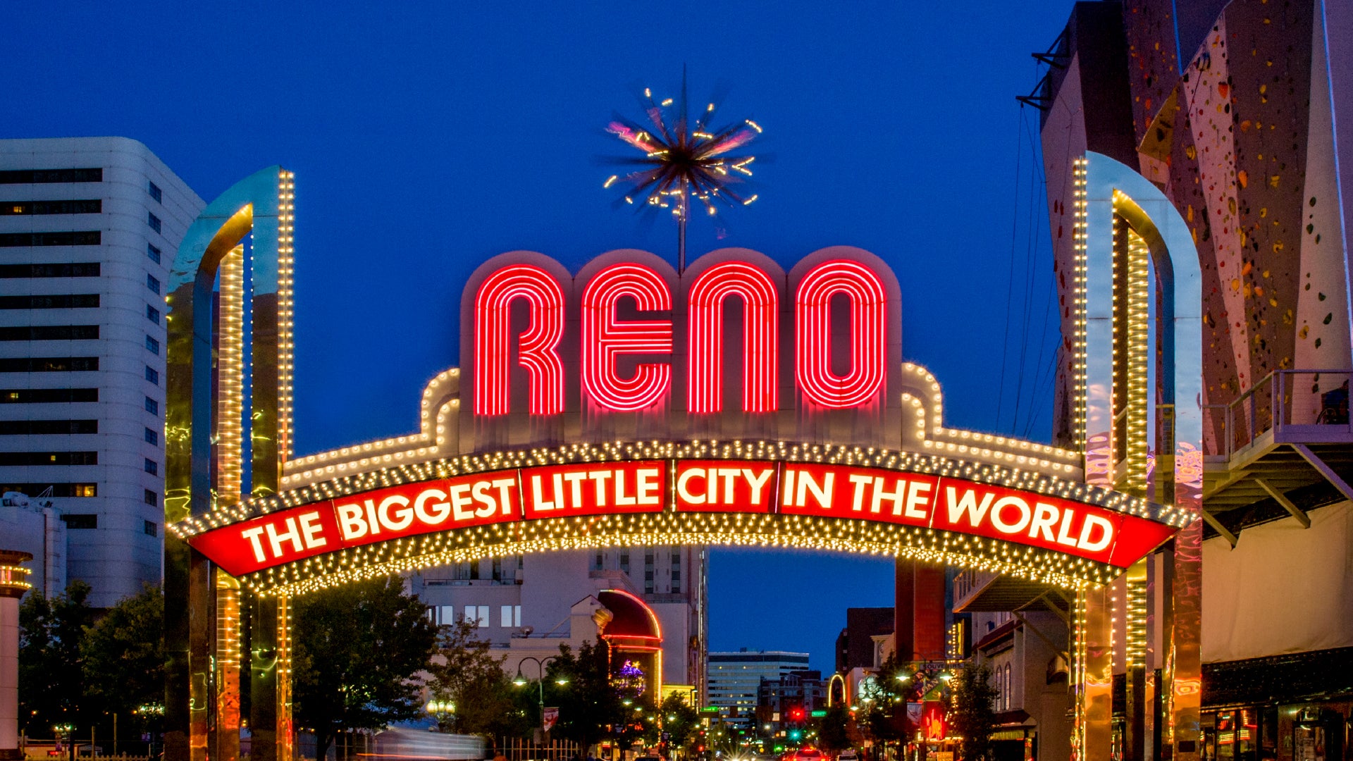 A vibrant nighttime scene featuring the illuminated Reno Arch in Reno, Nevada. The arch displays bold red neon letters spelling "RENO," with the slogan "THE BIGGEST LITTLE CITY IN THE WORLD" in white text on a red banner below. Golden lights outline the structure, creating a dazzling effect. A decorative starburst light fixture sits atop the arch. The surrounding cityscape includes modern buildings, neon signs, and a rock climbing wall on the right side. The deep blue evening sky provides a striking contras