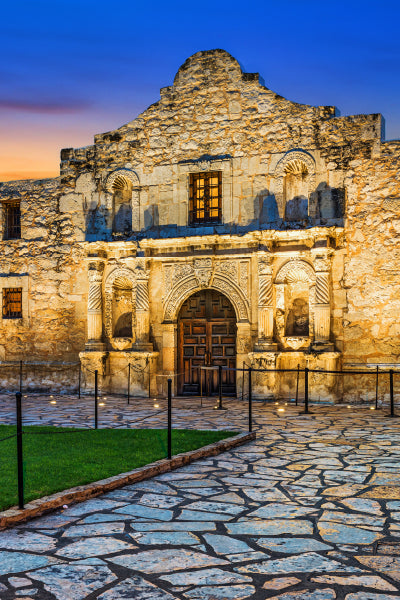 The historic Alamo mission building in San Antonio, Texas, illuminated at dusk with warm lights. The stone facade features an arched wooden door, detailed carvings, and a cobblestone pathway leading up to it, against a backdrop of a clear blue and orange evening sky.