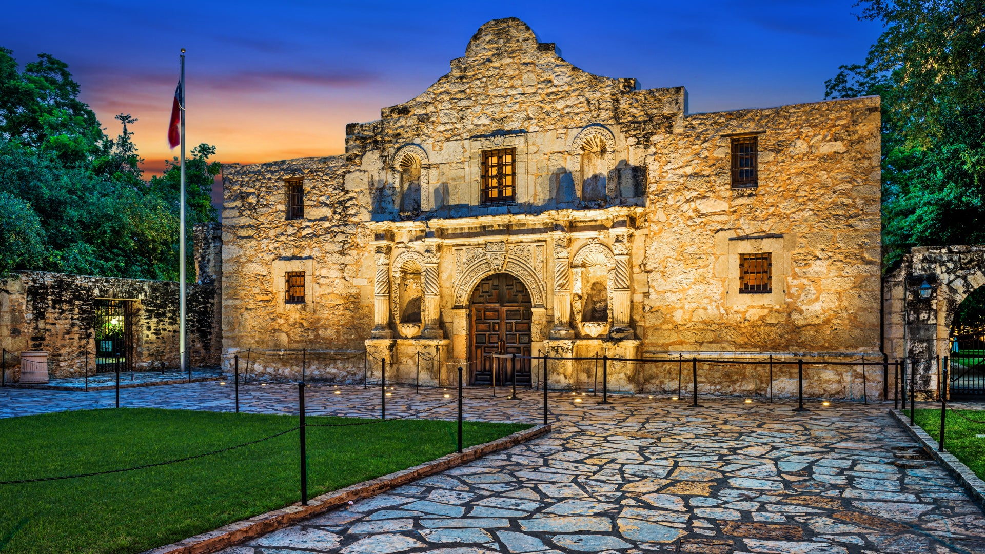 A historic stone building, the Alamo Mission in San Antonio, Texas, illuminated by warm golden lights at dusk. The sky displays a gradient of deep blue and orange hues. The textured limestone facade features ornate carvings around the wooden doorway, with small windows and decorative arches. A flagpole with a waving Texas flag stands to the left, while a stone courtyard with a pathway leads up to the entrance, bordered by green grass and low metal posts. Trees and stone walls surround the historic site, add